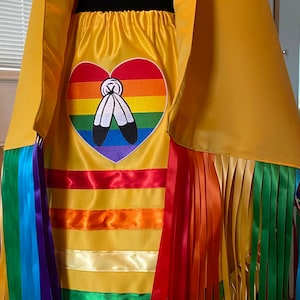 Könnte beinhalten: Ein gelbes Satinkleid mit Regenbogen-Bandfransen und einem Regenbogenherz mit Federdesign. Das Kleid ist im traditionellen Stil der amerikanischen Ureinwohner.