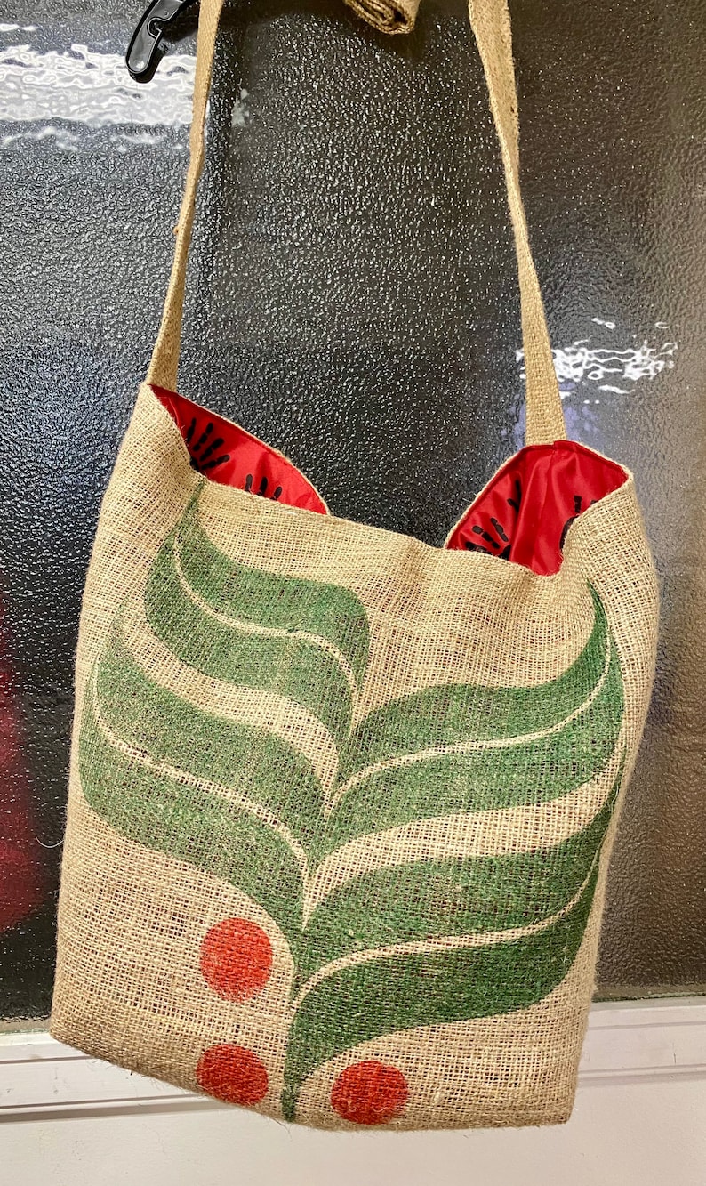 May include: A brown burlap tote bag with a green leaf design and red circles. The bag has a red lining and a brown strap.