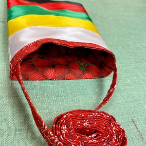 May include: A red and white fabric bag with a black, green, and yellow striped top. The bag has a red and white patterned strap.