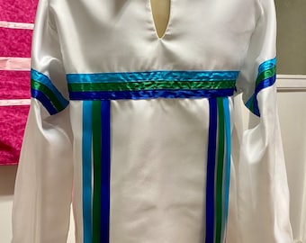 Puede incluir: Camisa blanca de manga larga con cuello en V y cintas decorativas azules y verdes. La camisa presenta detalles de cintas azules y verdes en el cuello, las mangas y la cintura. La camisa se exhibe en un maniquí.