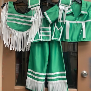 Op de afbeelding: Een groene en witte outfit met franjes. De outfit bestaat uit een vest, een shirt, een broek en een halsstuk.