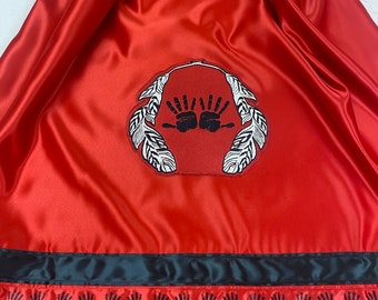 Peut inclure: Tissu de satin rouge avec un motif brodé noir de deux mains à l'intérieur d'un cercle de plumes. Le bord inférieur du tissu est orné d'un ruban noir avec un motif répétitif de mains noires.