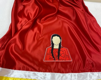 Può includere: Una gonna di raso rosso con un bordo bianco, giallo e nero. La gonna presenta un'immagine ricamata di una donna con lunghi capelli neri che indossa una camicia con motivi rossi e neri.