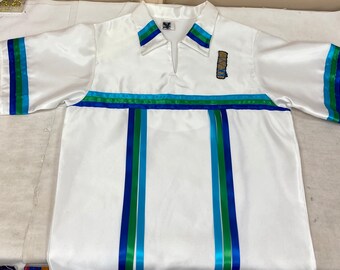 Puede incluir: Camisa blanca de manga corta con ribetes de cinta azul, verde y turquesa. La camisa tiene un cuello en V y un pequeño diseño bordado en la parte delantera.