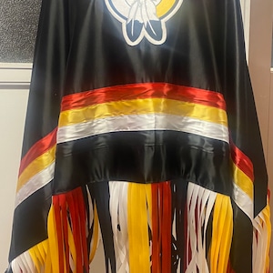 May include: Black ceremonial shawl with a circular emblem featuring a crosshair design and feathers. The shawl has horizontal bands of red, yellow, and white ribbon, and a fringe of colorful ribbons.