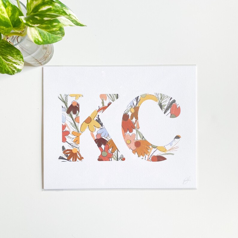 May include: A white print featuring the letters "KC" filled with a floral pattern in shades of yellow, orange, and brown. The artwork is on a white background, with a plant in a glass vase in the upper left corner.
