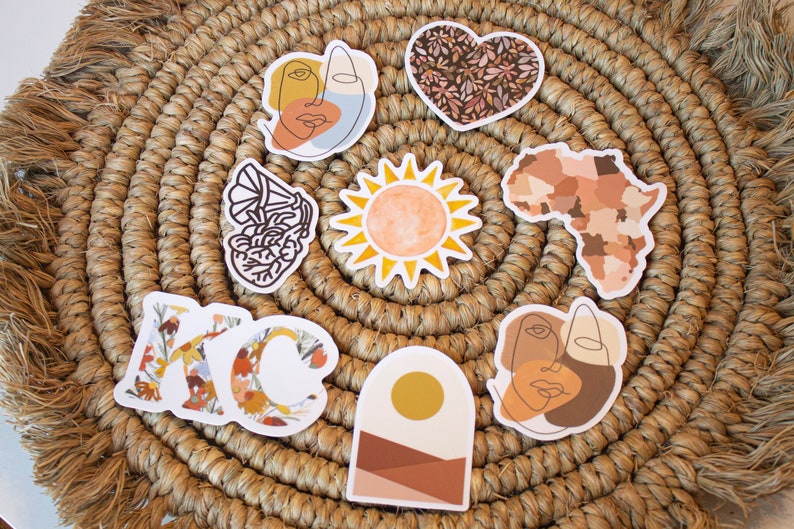 May include: A collection of decorative stickers with various designs, including a sun, a heart with floral patterns, an outline of Africa, and abstract face art. The stickers are arranged on a woven, circular placemat.