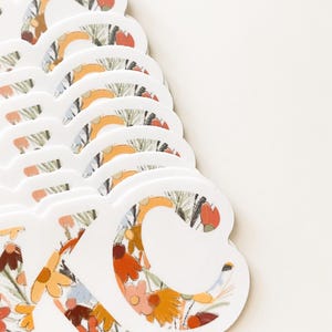 May include: A stack of white die-cut stickers with the letters "KC" in a floral pattern. The floral design features orange, yellow, and red flowers with green leaves. The stickers are arranged on a white background.