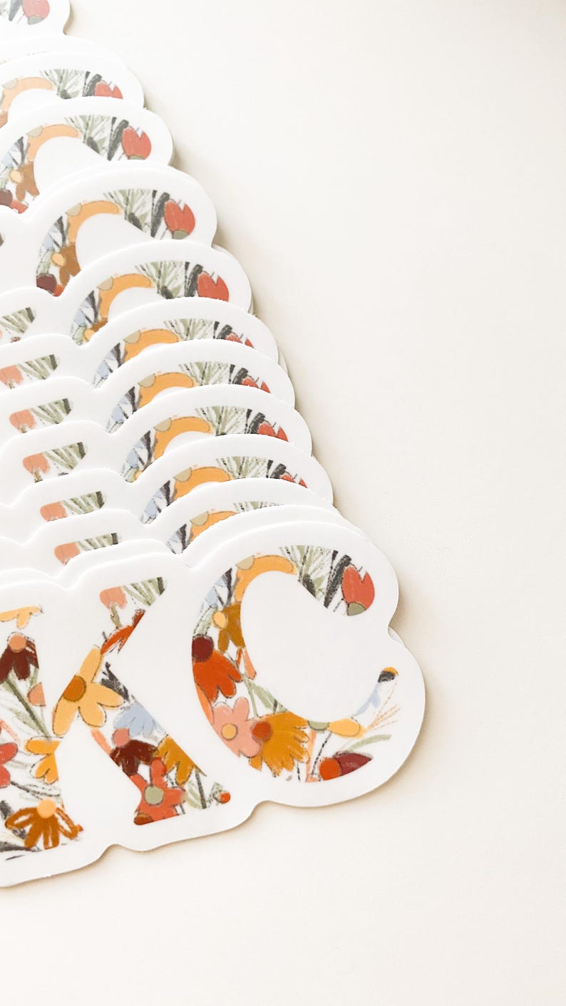 May include: A stack of white die-cut stickers with the letters "KC" in a floral pattern. The floral design features orange, yellow, and red flowers with green leaves. The stickers are arranged on a white background.