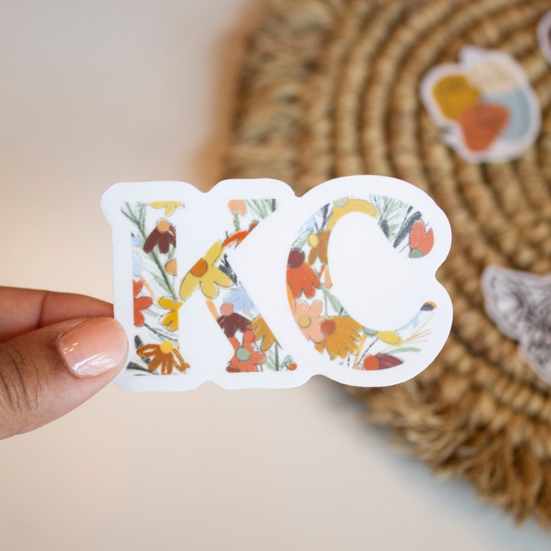 May include: A white sticker with the letters "KC" filled with a floral pattern in yellow, orange, and brown. The sticker is held by a hand, with a woven background visible.