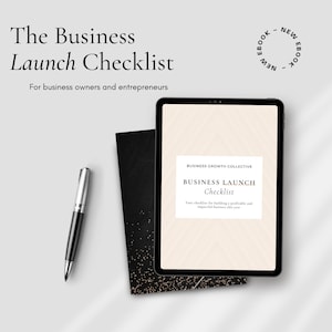 May include: A digital tablet displaying a business launch checklist ebook with the title "Business Launch Checklist" and the text "Your checklist for building a profitable and impactful business this year." The tablet is on a white surface with a black notebook and a silver pen.