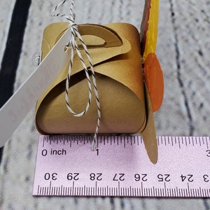 May include: A small brown paper box with a handle and a turkey design. The box is decorated with fall-colored leaves and is approximately 2.5 inches wide.
