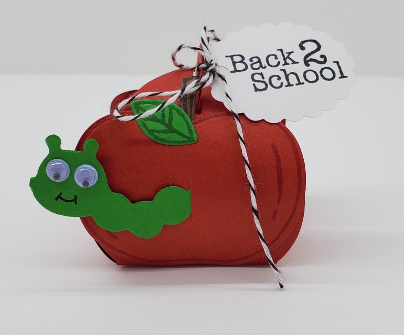 May include: A red paper apple with a green leaf and a green caterpillar with googly eyes. The apple has a white string tied around it with a white tag that says "Back 2 School".