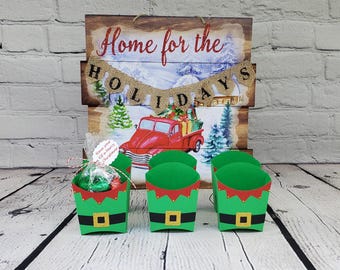 May include: Six green and red elf-themed treat boxes with a red and white "Home for the Holidays" sign in the background. The boxes have a white belt and a red and white elf hat.