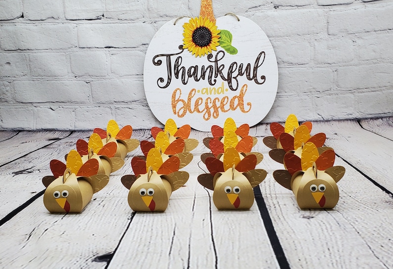 May include: A set of 12 brown paper turkey favor boxes with orange and yellow feathers. The boxes are arranged in two rows of six. The boxes are decorated with googly eyes and a red beak. A sign that says "Thankful and Blessed" with a sunflower is in the background.