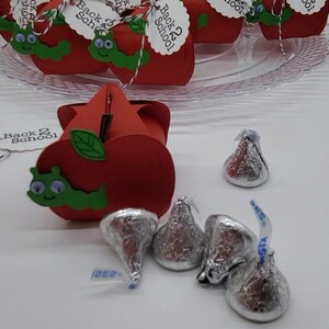 May include: Red apple-shaped boxes with green leaves and a green worm with googly eyes. The boxes have a tag that says "Back 2 School". There are silver foil-wrapped chocolate kisses on a white surface.