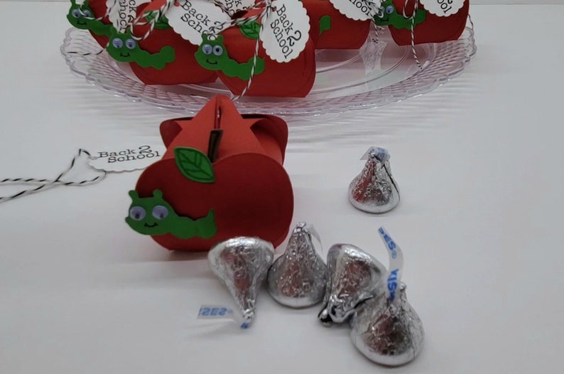 May include: Red apple-shaped boxes with green leaves and a green worm with googly eyes. The boxes have a tag that says "Back 2 School". There are silver foil-wrapped chocolate kisses on a white surface.