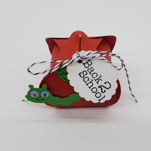 May include: Red and green paper craft box with a white tag that says "Back 2 School". The box is shaped like an apple and has a green worm on the front.