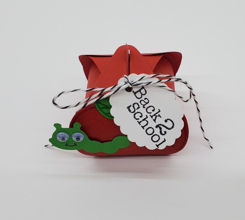 May include: Red and green paper craft box with a white tag that says "Back 2 School". The box is shaped like an apple and has a green worm on the front.