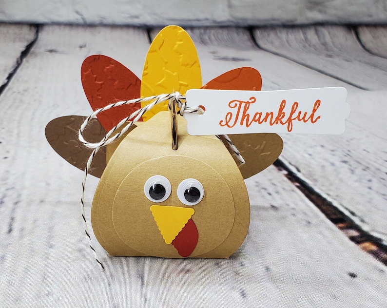 May include: A brown paper turkey-shaped box with googly eyes, a yellow beak, and a red wattle. The turkey has a white string tied around its neck with a white tag that says "Thankful".