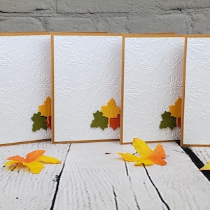 May include: Set of six blank greeting cards with a fall theme. Each card features a brown border and a white interior with a textured pattern. The cards are decorated with three layered paper leaves in shades of green, yellow, and orange.