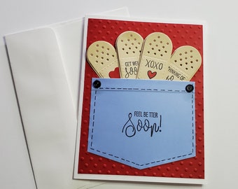 May include: A red card with white polka dots and a blue pocket. The pocket has four band-aid shaped cards sticking out of it. The band-aids have the text "Get Well Soon", "XOXO", and "Thinking of You" on them. The pocket has the text "Feel Better Soon!" on it.