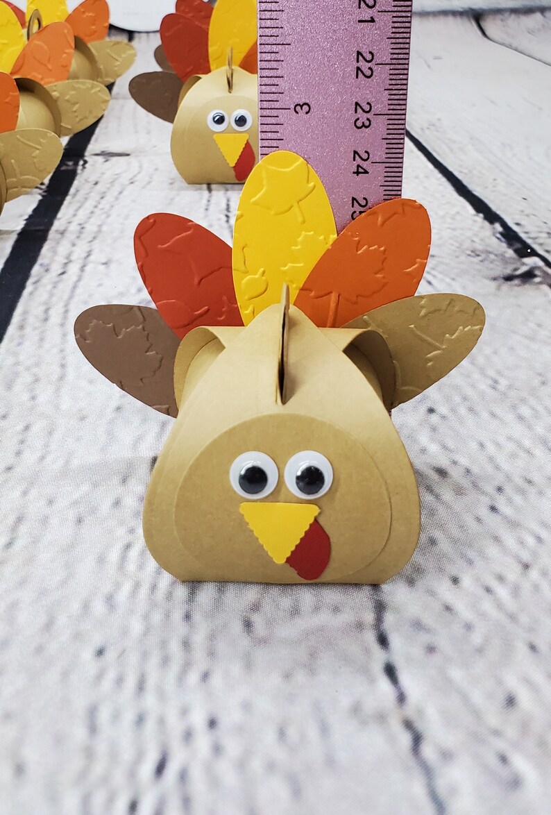 May include: A brown paper turkey favor box with a yellow beak, red wattle, and googly eyes. The turkey has a brown, orange, and yellow patterned tail.