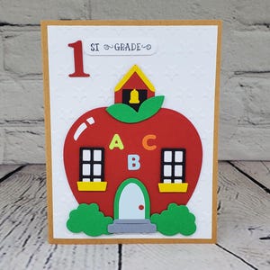 May include: A red apple shaped card with a schoolhouse on top. The schoolhouse has a yellow roof, a red door, and a bell. The apple has windows and the letters A, B, and C on the front. The card says "1st Grade" on the top.