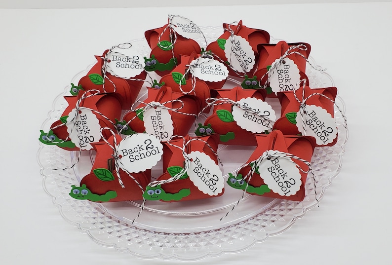 May include: A collection of red apple-shaped treat boxes with green leaves and a cartoon caterpillar on the front. Each box has a white tag tied with twine that says "Back 2 School".