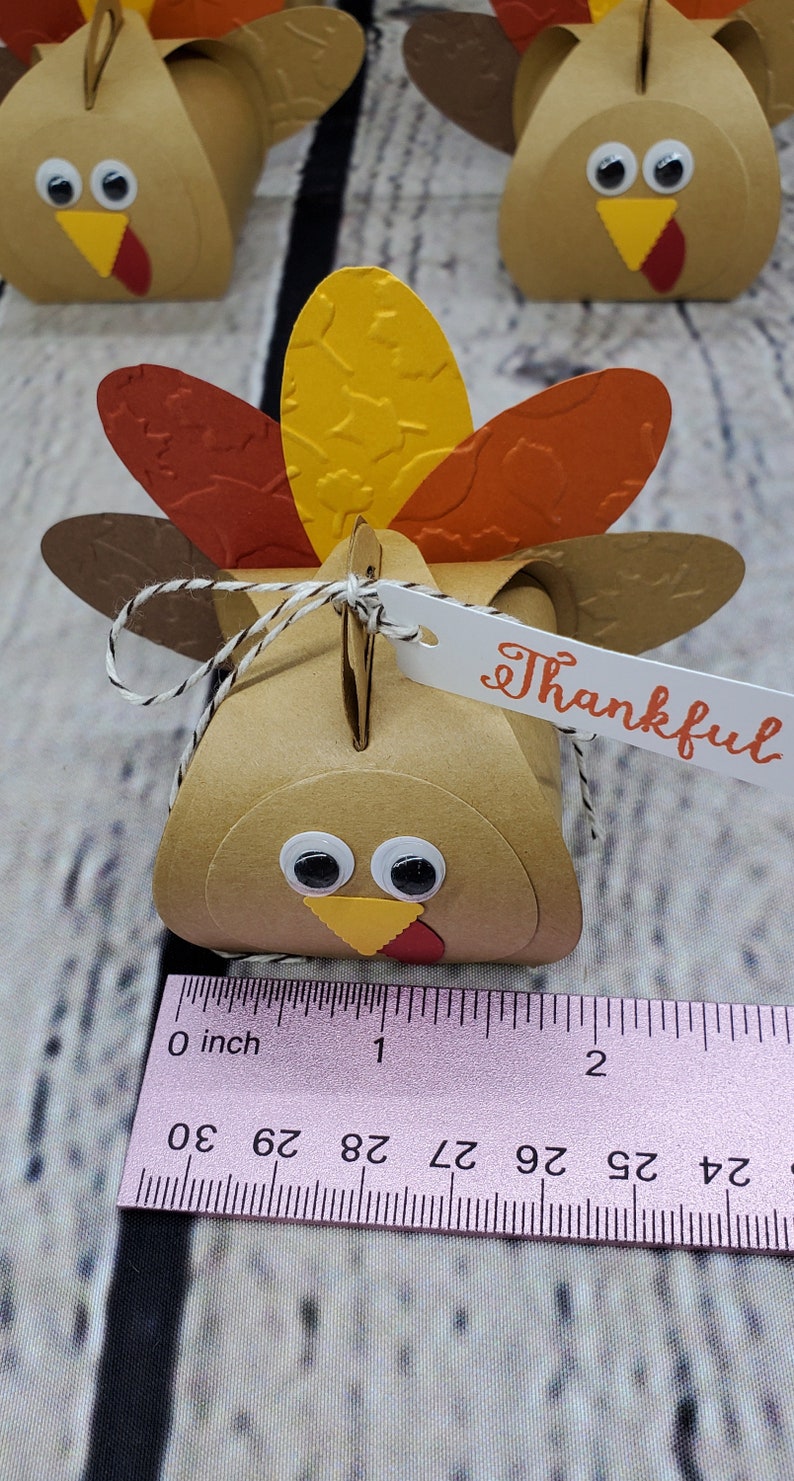 May include: A brown paper turkey favor box with a yellow beak, red wattle, and googly eyes. The box has a tag that says "Thankful".