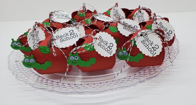 May include: A platter of red apple-shaped boxes decorated with green worms and leaves. Each box has a white tag with the text "Back 2 School" in black.
