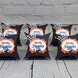 May include: Six black pillow boxes with orange and white spiderweb designs. Each box has a white circle with the text "Spooky Sweets" in black. A black spider silhouette is in the center of each circle.
