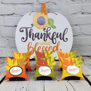 May include: A white pumpkin-shaped sign with the words "Thankful and Blessed" and a sunflower decoration. Three decorative gift boxes in orange, green, and yellow, each with a leaf design and the words "thankful for you."