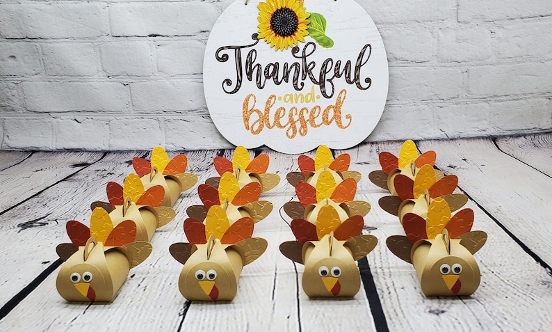 May include: A row of 12 brown paper turkey favor boxes with orange, yellow, and brown feathers. The boxes are arranged in two rows of six. The boxes are decorated with googly eyes and orange beaks. The boxes are sitting on a white wooden surface.