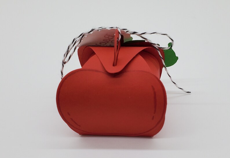 May include: Red apple-shaped paper box with a green leaf and a white tag with black text that says "Best Teacher Ever". The box is tied with a white and black twine.