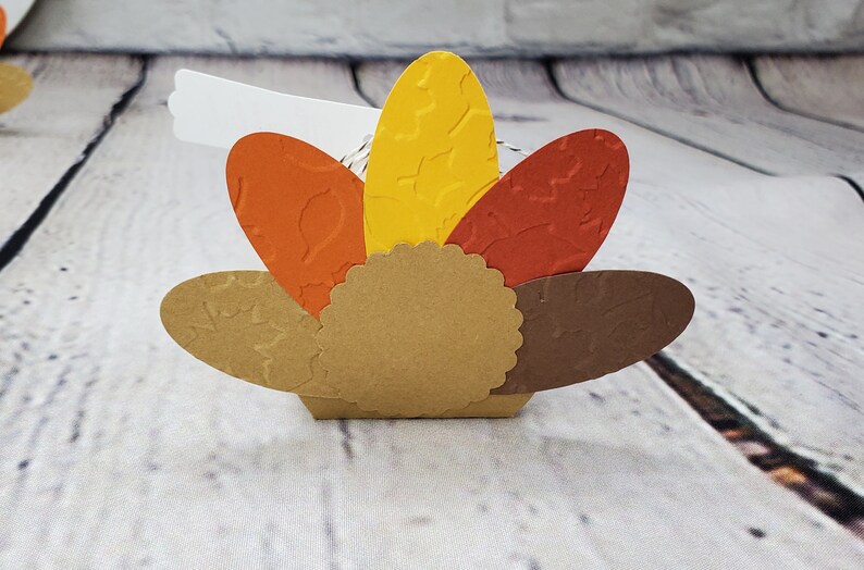 May include: A paper craft turkey favor box with a brown body, orange and yellow feathers, and a white tag.