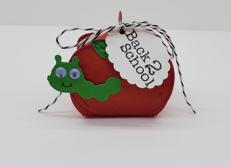 May include: A red apple-shaped box with a green worm and googly eyes. The box has a white tag that says "Back 2 School".