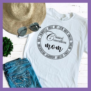 Puede incluir: Camiseta blanca con un dise&ntilde;o circular negro que dice "Classical Conversations mom" con las palabras "Biblia, Tin Whistle, M&uacute;sica, Arte, Lat&iacute;n, Matem&aacute;ticas, Ingl&eacute;s, Cronolog&iacute;a, Ciencia, Historia, Geograf&iacute;a, Presentaciones" alrededor del c&iacute;rculo.