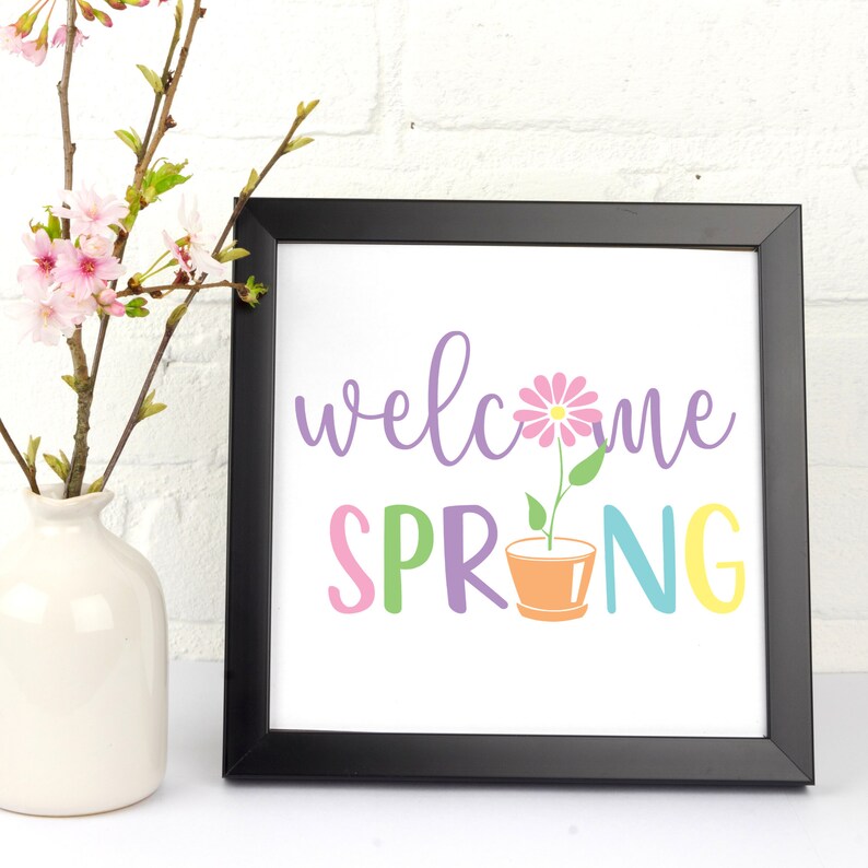 K&ouml;nnte beinhalten: Ein schwarzer gerahmter Druck mit dem Text "welcome spring" in bunten Buchstaben. Eine rosa Blume in einem Topf befindet sich mittig zwischen den W&ouml;rtern.