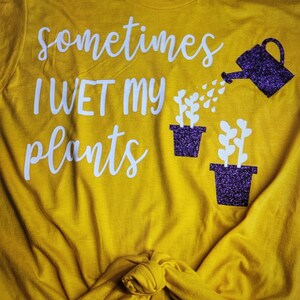 May include: A yellow t-shirt with the text "sometimes I WET my plants" in white lettering. A purple glitter watering can is pouring water onto two purple glitter potted plants.