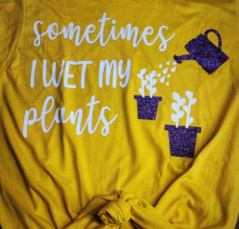 May include: A yellow t-shirt with the text "sometimes I WET my plants" in white lettering. A purple glitter watering can is pouring water onto two purple glitter potted plants.