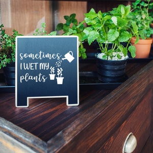 May include: A black chalkboard sign with white text that reads "sometimes I WET MY plants" and a graphic of a watering can pouring water on two potted plants. The sign is sitting on a wooden surface with potted plants in the background.