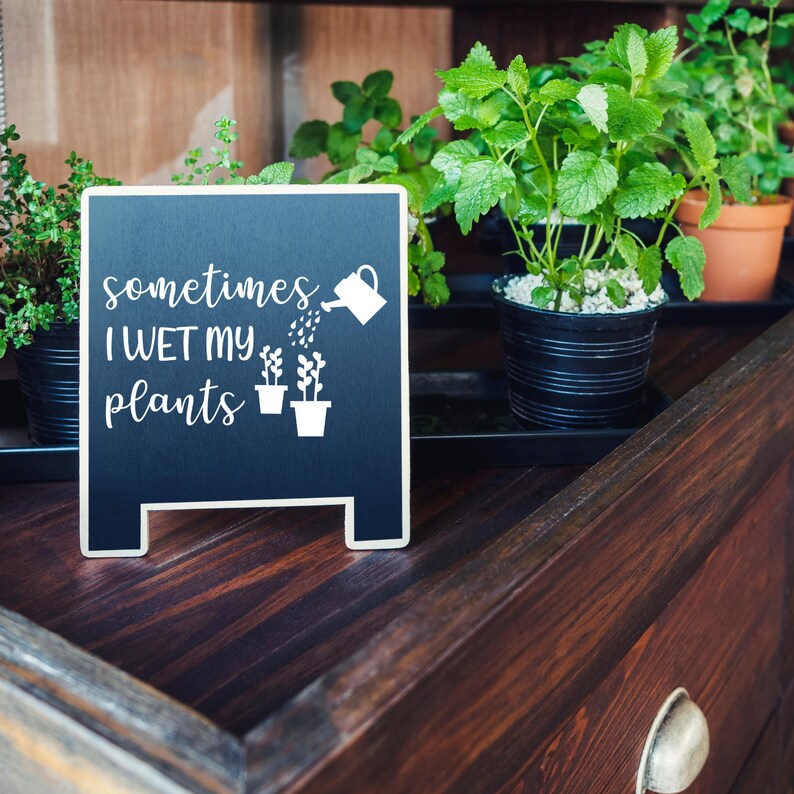 May include: A black chalkboard sign with white text that reads "sometimes I WET MY plants" and a graphic of a watering can pouring water on two potted plants. The sign is sitting on a wooden surface with potted plants in the background.