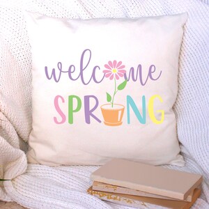 K&ouml;nnte beinhalten: Ein wei&szlig;es Kissen mit dem Text "welcome spring" in Lila, Rosa, Gr&uuml;n, Blau und Gelb. Eine rosa Blume mit einem gr&uuml;nen Stiel w&auml;chst in der Mitte des Textes aus einem braunen Topf.