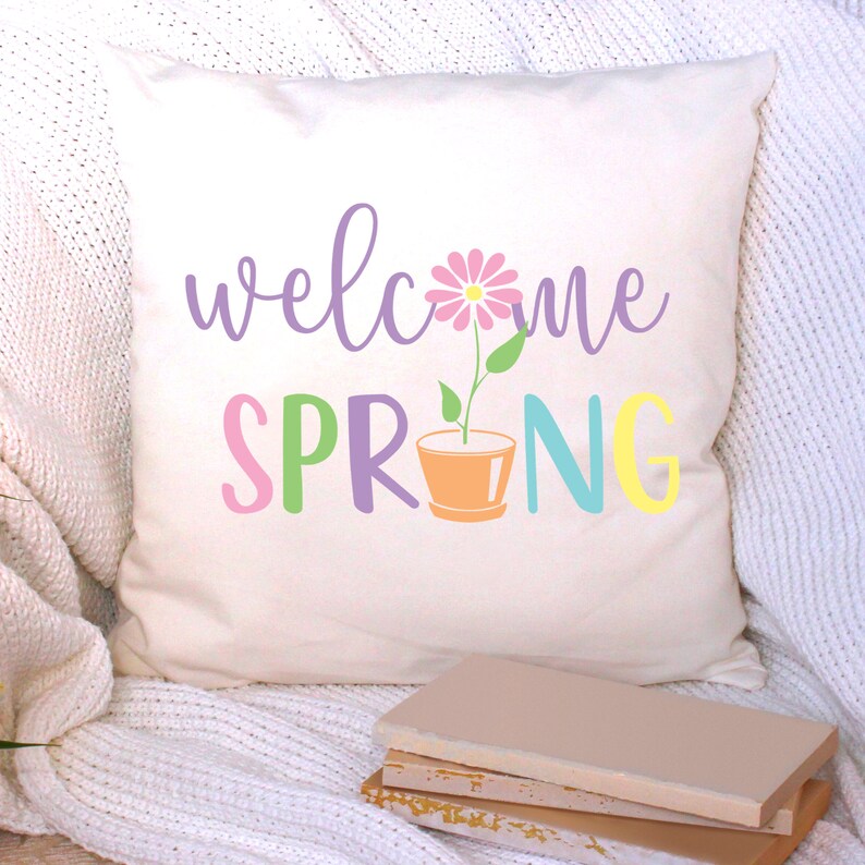 K&ouml;nnte beinhalten: Ein wei&szlig;es Kissen mit dem Text "welcome spring" in Lila, Rosa, Gr&uuml;n, Blau und Gelb. Eine rosa Blume mit einem gr&uuml;nen Stiel w&auml;chst in der Mitte des Textes aus einem braunen Topf.