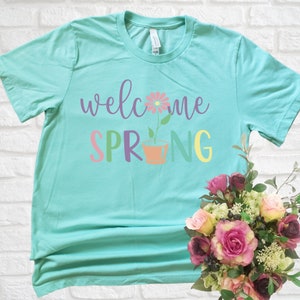 K&ouml;nnte beinhalten: Ein t&uuml;rkisfarbenes T-Shirt mit dem Text "welcome spring" in bunten Buchstaben. Eine rosa Blume mit einem gr&uuml;nen Stiel befindet sich in einem Terrakotta-Topf zwischen den W&ouml;rtern "spring".
