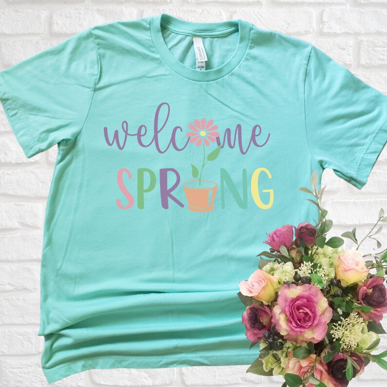 K&ouml;nnte beinhalten: Ein t&uuml;rkisfarbenes T-Shirt mit dem Text "welcome spring" in bunten Buchstaben. Eine rosa Blume mit einem gr&uuml;nen Stiel befindet sich in einem Terrakotta-Topf zwischen den W&ouml;rtern "spring".