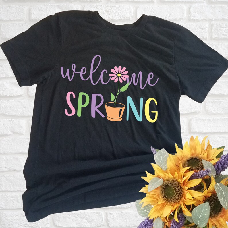 K&ouml;nnte beinhalten: Schwarzes T-Shirt mit dem Text "welcome spring" in bunten Buchstaben. Eine rosa Blume in einem braunen Topf befindet sich in der Mitte des Textes.