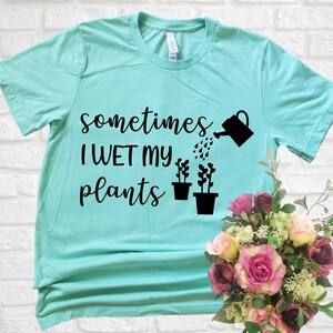 May include: A turquoise blue t-shirt with the text "sometimes I WET my plants" in black lettering. A black watering can is pouring water onto three black potted plants.