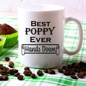 May include: White ceramic coffee mug with black text that reads "Best Poppy Ever Hands Down". The mug is sitting on a green and white checkered tablecloth with coffee beans scattered around it.
